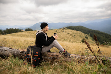 Naklejka na ściany i meble Young woman with a thermos cup resting on a fallen tree in the mountains. Freedom, food, travel and vacation concept, outdoor activities.