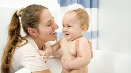 Portrait of smiling baby boy covred in soap foam and mother washing him in bathroom