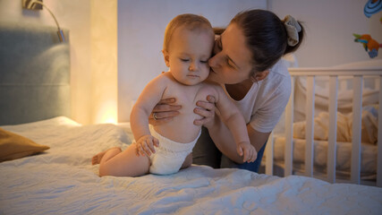 Young smiling mother dressing and putting on diapers on her baby son in bed at night. Concept of children hygiene, healthcare and family care at home