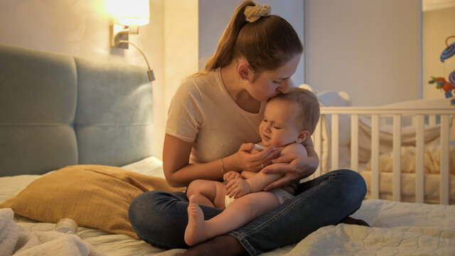 Young loving mother kissing her baby son while measuring his temperature with thermometer