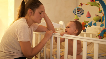 Tired sleepy mother rocking crib of her baby crying and refusing to sleep at night. Concept of...