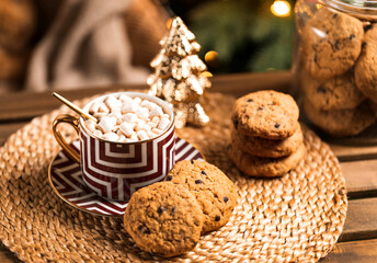 A cup of cocoa with marshmallows and american cookies on wooden coffee table with Christmas or New Year decorations over defocused Christmas tree with fairy lights and christmas tree toys at home.