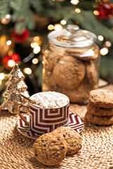 A cup of cocoa with marshmallows and american cookies on wooden coffee table with Christmas or New Year decorations over defocused Christmas tree with fairy lights and christmas tree toys at home.