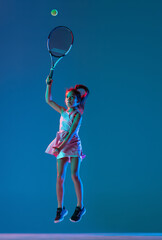 Portrait of little girl, beginner tennis player playing tennis isolated on blue studio background in neon light. Sport, study, childhood concept