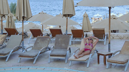 A middle-aged woman in a bikini lies on a sun lounger by the pool by the sea. She is enjoying her time at the hotel with the palm trees in the background.