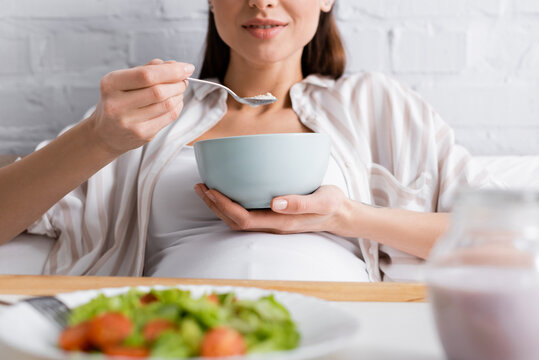 cropped view of happy pregnant woman eating oatmeal near tray with salad.