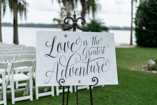 White Wooden sign with Love is the greatest adventure words being used for a wedding ceremony in the outdoors Wedding decoration details White Rows of chairs background