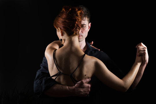 Young couple - a man and a woman wearing black dancing in the dark; the lady stands with her back to the camera.