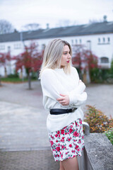 Freezing cold european white young woman with blonde hair in white sweater in floral skirt in a city