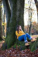 Young european white woman in yellow sweater sitting near tree trunk in sunny autumn forest