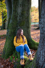 Smiling young european white woman in yellow sweater sitting near tree trunk in sunny autumn forest