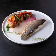 Fish with potatoes and stewed vegetables on a white plate on a dark background, shot from the side