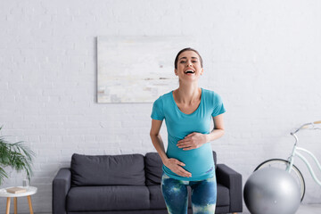 Fototapeta na wymiar cheerful pregnant sportswoman touching belly and laughing in living room.