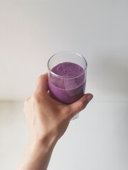Blueberry velvet violet smoothie in a transparent glass in women's hands on gray background with long shadows, top view. Milk Shakes. Healthy breakfast. Green living and eco-friendly products