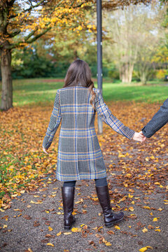 Turned back young woman in trench coat in autumn park holding someone hand