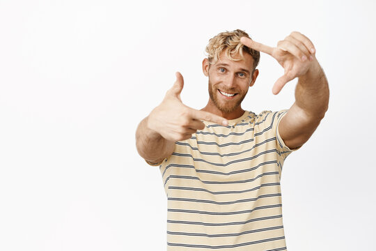 Image of smiling blond guy stretching out hands frame and looking happy, picturing moment, taking photo, standing over white background