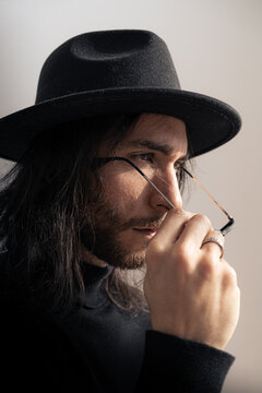 Hombre de pelo largo con gafas de sol, sombrero, sueter de cuello vuelto, barba, y raza cauc&aacute;sica, sujetando sus gafas de sol con la mano