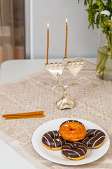Lighted Hanukkah with one candle and shamash on the first day of Hanukkah on the festive table next to donuts and a vase of flowers.