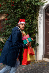 Christmas shopping time. Young man with Santa Claus hat has a surprised expression and brings his newly purchased gifts.