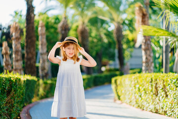 little girl in a white dress and a straw hat on the path in a tropical hotel