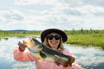 Woman fishing on a lake holding big bass catch. Lady holding largemouth bass catch with sun shinning. Text space. - obrazy, fototapety, plakaty