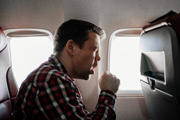 a man in a plaid shirt coughs on the plane in a window seat. 