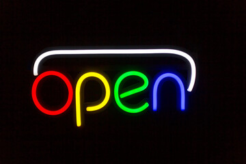 A neon sign that reads "Open" hangs on a black background. Neon Light "Open" Sign hanging above building at night. Glowing open neon sign in a window.