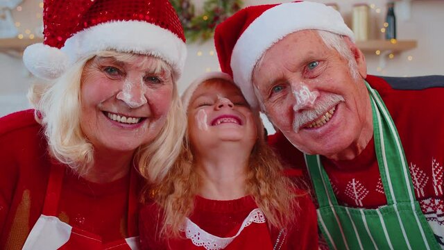 POV shot of mature grandparents couple family with granddaughter kid smeared face with flour taking selfie on mobile phone during online video call at home Christmas kitchen. Celebrating Xmas holidays