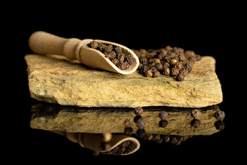 Lot of whole spicy black pepper in a wooden scoop with natural flat rock isolated on black glass