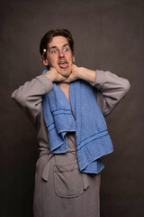 Young handsome tall slim white man with brown hair choking himself with towel on shoulders in grey bathrobe on grey background