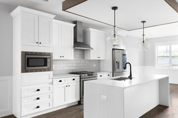 A renovated white kitchen with glass pendant lights hanging above the waterfall granite island, stainless steel appliances, and hardwood floors.