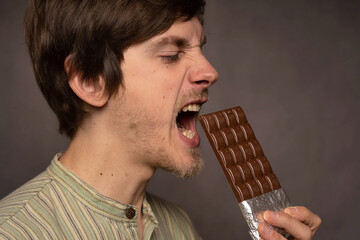 Young handsome tall slim white man with brown hair about to bite chocolate bar in striped shirt on grey background