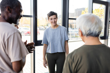 Smiling senior woman standing near interracial friends in gym.