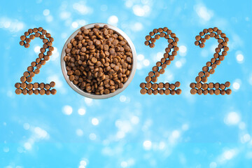 Date 2022 made of dry food and a round metal bowl full of crunchy dry food for dogs or cats on a blue background with bokeh