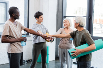 Elderly interracial people holding fitness mats and hands in gym.
