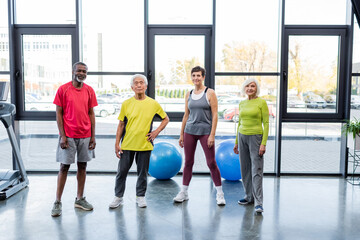 Fototapeta premium Positive multiethnic senior people looking at camera near fitness balls and treadmill in gym.