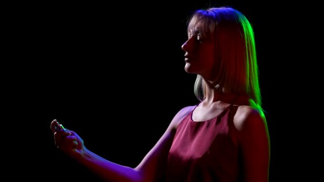 Side view of a silhouette of young pretty blonde sprinkling perfume on herself from a spray bottle. Portrait of a cute girl posing over black studio background with neon lights. Close up. Slow motion.