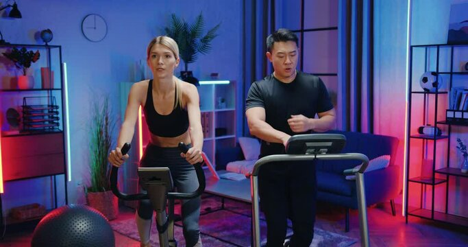 Attractive Active Sporty Motivated Fit Woman And Asian Man Working Out Together At Home,she Twisting Pedals On Stationary Bike,he Running On Treadmill