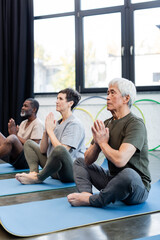 Elderly asian man sitting in yoga pose near multiethnic friends in sports center.
