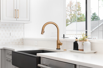 A beautiful sink in a remodeled modern farmhouse kitchen with a gold faucet, black apron or farmhouse sink, white granite, and a tiled backsplash. - Powered by Adobe