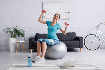 Fototapeta na wymiar happy pregnant woman exercising on fitness ball with dumbbells near laptop.
