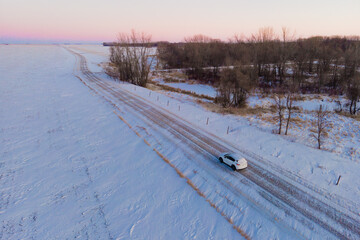 Aerial photo of a white SUV driving down a snowy road in rural North Dakota. Snow is blowing across the road and barren, brown vegetation surrounds the road. Beautiful sunset. Winter.