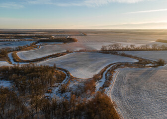 Aerial view of winter trees, frozen, snowy farmland and cultivated fields in rural North Dakota. Winter, sunset, sun flares. Vast expanse with no one.