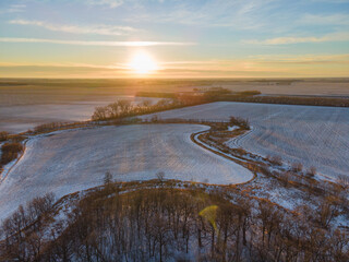 Aerial view of winter trees, frozen, snowy farmland and cultivated fields in rural North Dakota. Winter, sunset, sun shining low in sky. Vast expanse with no one.