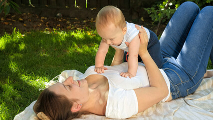 Smiling young mother lying on grass with her little baby son and smiling at him. Parenting, family, children development, and fun outdoors in nature.