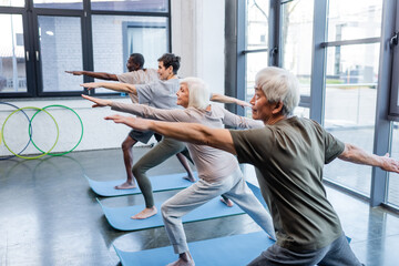 Senior woman in warrior pose practicing yoga near multiethnic people in gym.