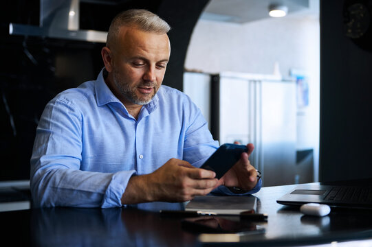 Attractive confident middle aged businessman, successful entrepreneur in blue casual shirt smiles using mobile phone sitting at table at stylish home interior