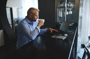Successful business man in business casual attire drinking coffee in the morning, sitting at a table with laptop and digital gadgets before getting ready to go at work