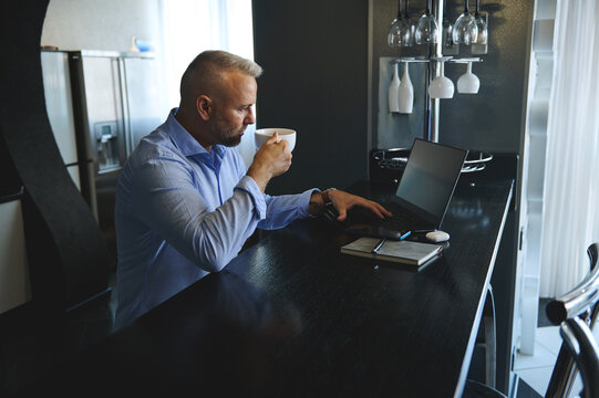 Successful Business Man In Business Casual Attire Drinking Coffee In The Morning, Sitting At A Table With Laptop And Digital Gadgets Before Getting Ready To Go At Work