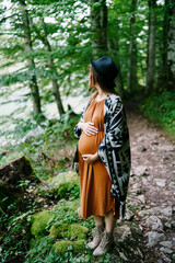 Pregnant woman stands on the path in the park hugging her belly with her hands